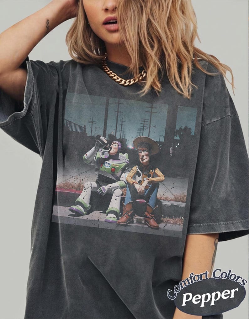 May include: A charcoal grey short-sleeve t-shirt featuring a graphic print of Buzz Lightyear and Woody sitting on a curb. The shirt has a washed-out, vintage appearance. The text "Comfort Colors Pepper" is printed in the bottom right corner.