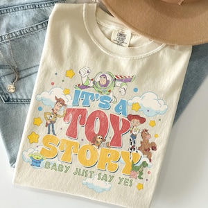 Peut inclure: Un t-shirt &agrave; manches courtes de couleur cr&egrave;me avec une impression graphique des personnages de Toy Story, dont Buzz l'&Eacute;clair, Woody, Jessie, Pile-Poil, Zigzag, Rex et un Alien. Le texte indique IT'S A TOY STORY BABY JUST SAY YES en lettres color&eacute;es.