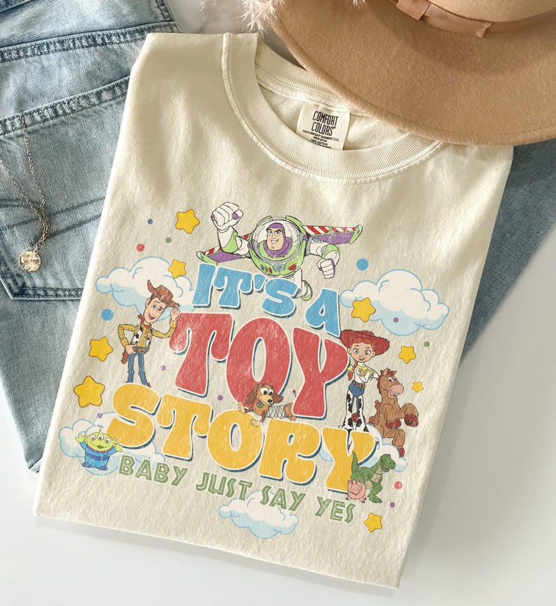 Peut inclure: Un t-shirt &agrave; manches courtes de couleur cr&egrave;me avec une impression graphique des personnages de Toy Story, dont Buzz l'&Eacute;clair, Woody, Jessie, Pile-Poil, Zigzag, Rex et un Alien. Le texte indique IT'S A TOY STORY BABY JUST SAY YES en lettres color&eacute;es.