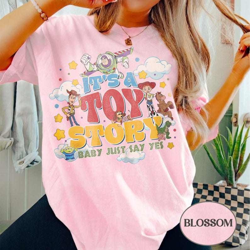 Peut inclure: Un t-shirt rose clair &agrave; manches courtes avec une impression graphique color&eacute;e des personnages de Toy Story, dont Buzz l'&Eacute;clair, Woody et Jessie. Le texte indique IT'S A TOY STORY BABY JUST SAY YES dans des polices r&eacute;tro bleues, rouges et jaunes.
