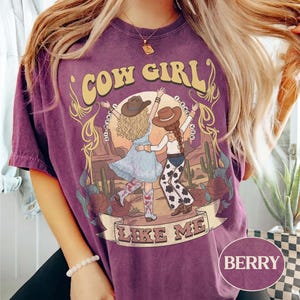 Puede incluir: Una camiseta de manga corta color baya con un estampado gr&aacute;fico de dos figuras con sombreros de vaquero bailando en un paisaje des&eacute;rtico. El texto dice COW GIRL y LIKE ME, con la frase I KNEW YOU rodeando a las figuras. Incluye cactus y rosas.