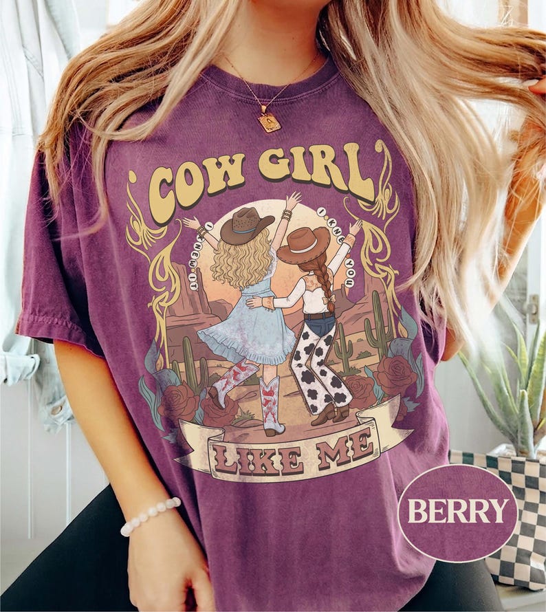 Puede incluir: Una camiseta de manga corta color baya con un estampado gr&aacute;fico de dos figuras con sombreros de vaquero bailando en un paisaje des&eacute;rtico. El texto dice COW GIRL y LIKE ME, con la frase I KNEW YOU rodeando a las figuras. Incluye cactus y rosas.