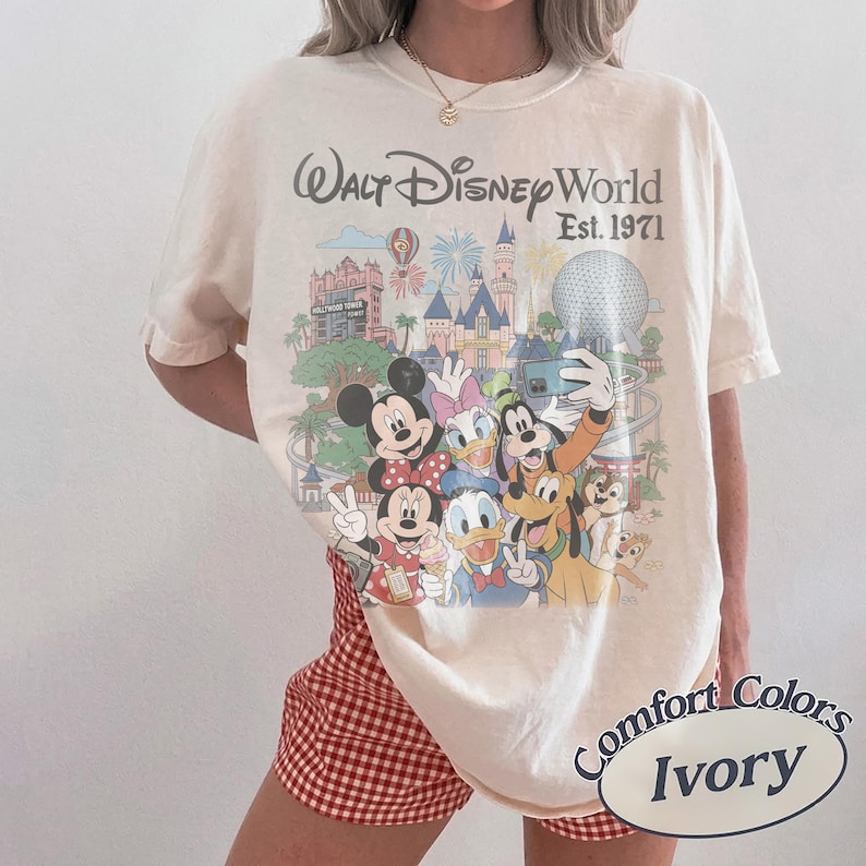 May include: An ivory short-sleeve t-shirt featuring a colorful graphic of Walt Disney World characters including Mickey Mouse, Minnie Mouse, Donald Duck, Daisy Duck, Goofy, Pluto, and Chip 'n Dale with the text Walt Disney World Est. 1971.
