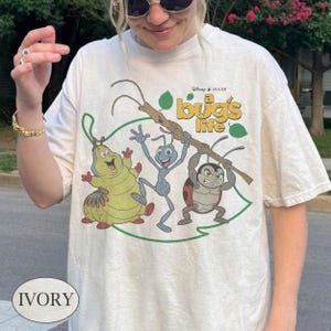 Puede incluir: Una camiseta de color marfil con un estampado gráfico de los personajes de la película A Bug's Life, que incluye una oruga amarilla, una hormiga azul y una mariquita sosteniendo un palo. El texto A Bug's Life está impreso en amarillo.