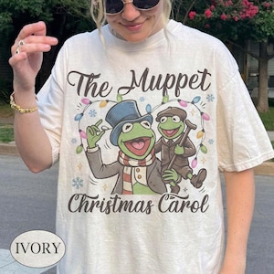May include: An ivory t-shirt featuring a graphic of Kermit the Frog in a top hat and scarf alongside another frog character holding a cane. The text reads The Muppet Christmas Carol, surrounded by a string of colorful holiday lights and snowflakes.