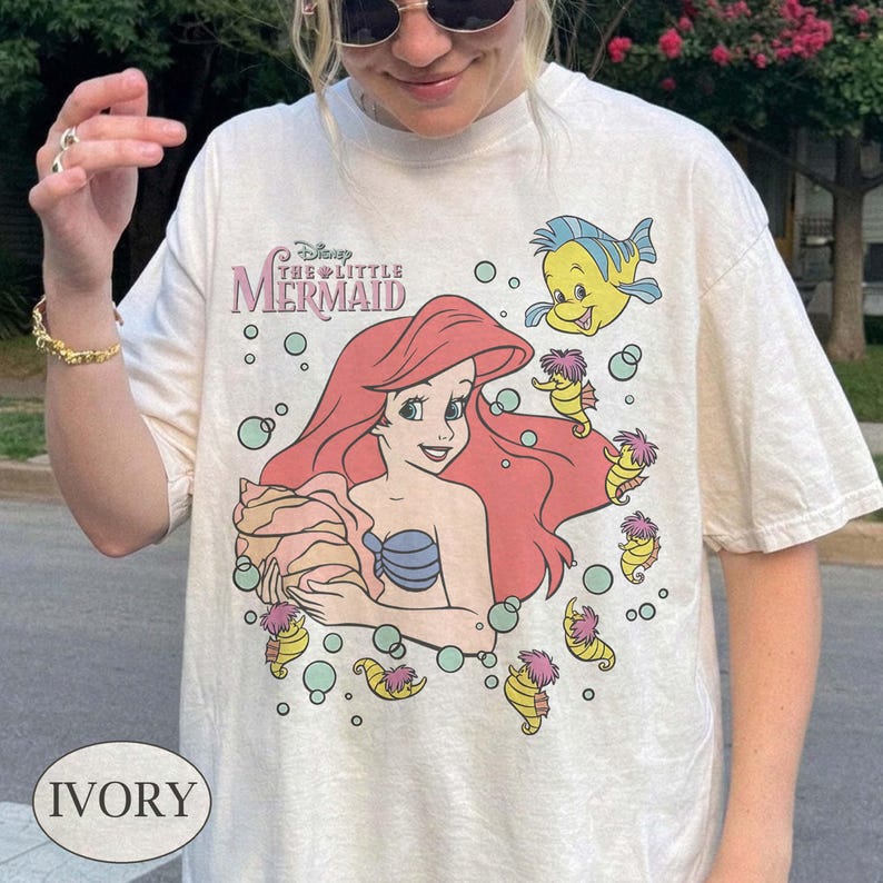 May include: Ivory t-shirt with a graphic of Ariel from Disney's The Little Mermaid. The design includes Ariel with red hair, a blue seashell bra, and a seashell. The shirt also features Flounder and seahorses.