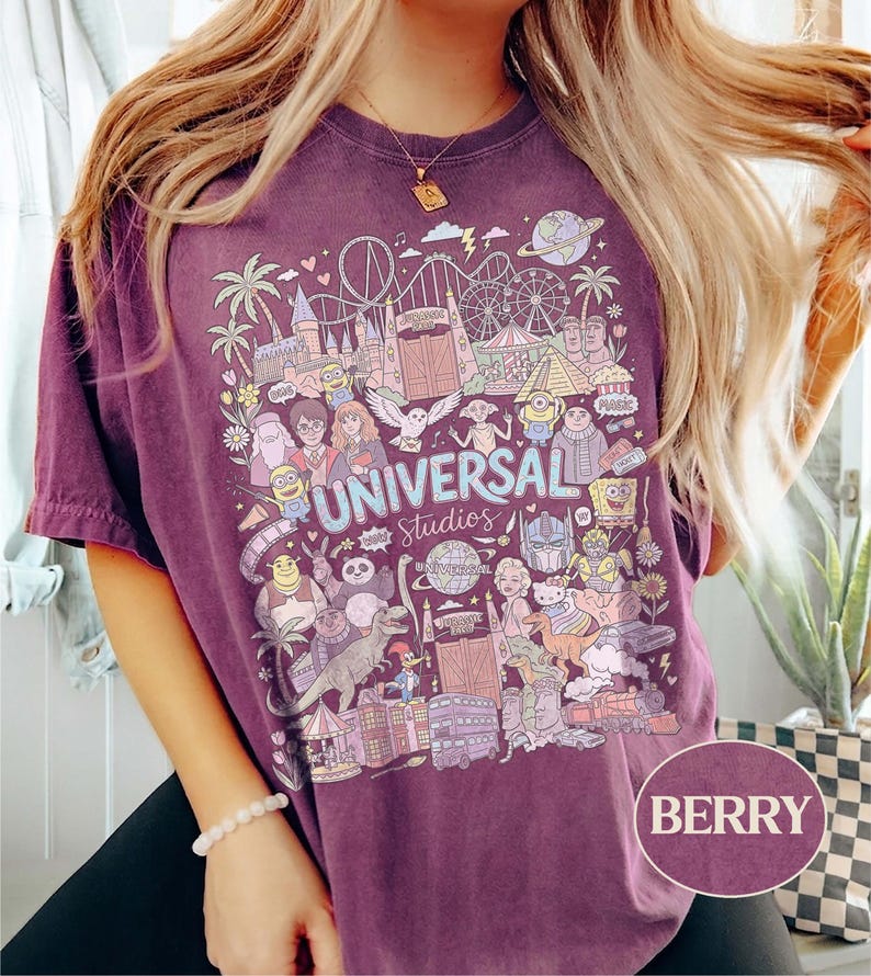 K&ouml;nnte beinhalten: Beerenfarbenes T-Shirt mit einer farbenfrohen Grafik von Universal Studios-Charakteren und -Attraktionen. Das Design enth&auml;lt den Text "UNIVERSAL Studios" und verschiedene Cartoon-Figuren. Das Shirt hat kurze &Auml;rmel und eine lockere Passform.