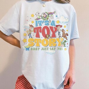 Peut inclure: Un t-shirt bleu clair avec une impression graphique color&eacute;e des personnages de Toy Story, dont Buzz l'&Eacute;clair, Woody et Jessie. Le texte indique IT'S A TOY STORY BABY JUST SAY YES, entour&eacute; d'&eacute;toiles jaunes et de nuages blancs.