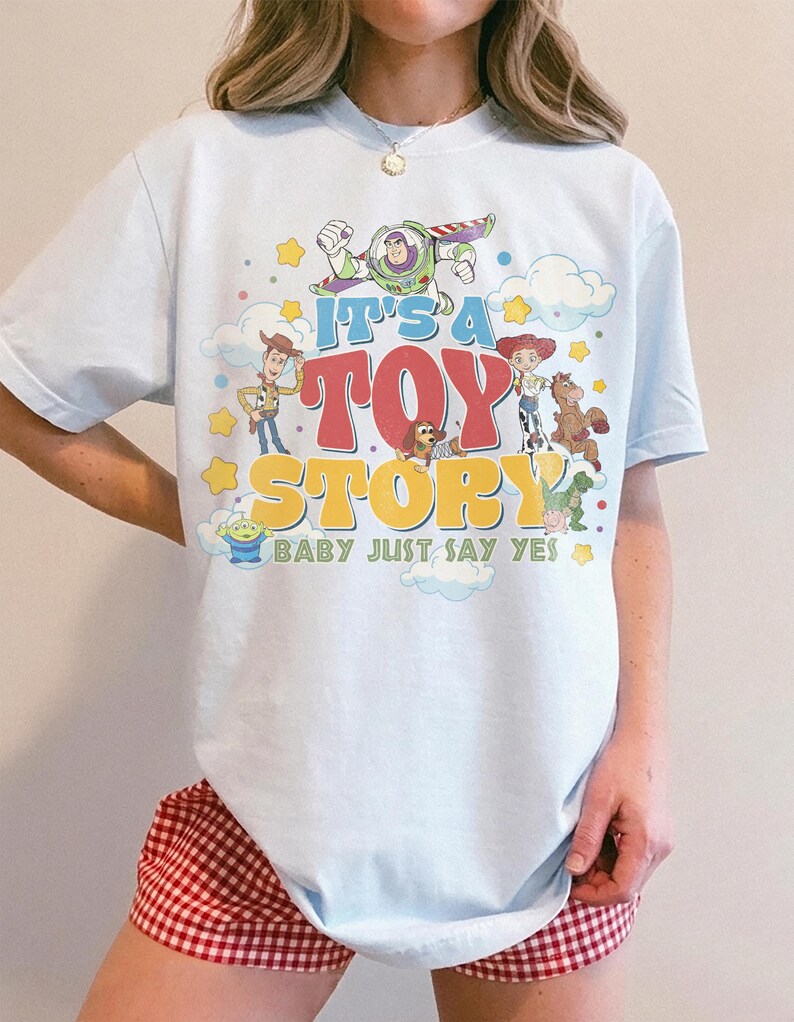 Peut inclure: Un t-shirt bleu clair avec une impression graphique color&eacute;e des personnages de Toy Story, dont Buzz l'&Eacute;clair, Woody et Jessie. Le texte indique IT'S A TOY STORY BABY JUST SAY YES, entour&eacute; d'&eacute;toiles jaunes et de nuages blancs.