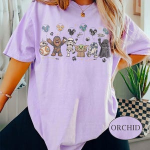 May include: A light purple t-shirt featuring a graphic of Star Wars characters holding balloons. The characters include BB-8, Chewbacca, a Stormtrooper, Grogu, R2-D2, and Darth Vader. The word "ORCHID" is printed on a white oval.