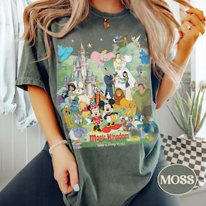 May include: A moss green t-shirt featuring a colorful graphic of Disney characters and the text "Magic Kingdom Walt Disney World". The design includes Mickey and Minnie Mouse, princesses, and other beloved characters.