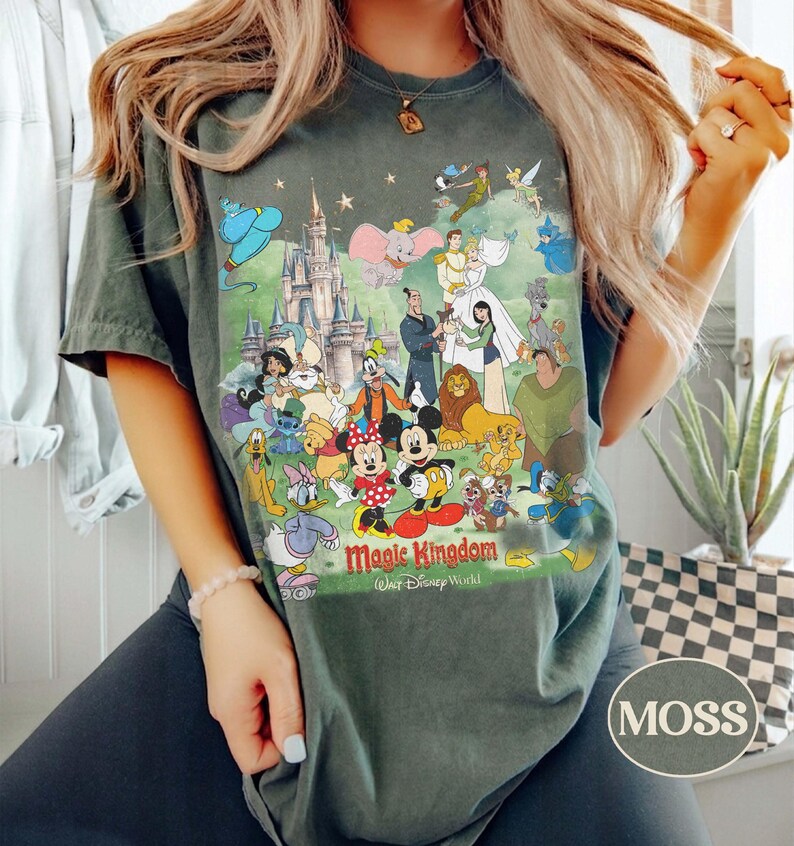 May include: A moss green t-shirt featuring a colorful graphic of Disney characters and the text "Magic Kingdom Walt Disney World". The design includes Mickey and Minnie Mouse, princesses, and other beloved characters.
