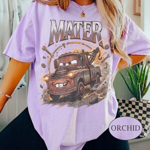 May include: A light purple t-shirt featuring a graphic print of a brown tow truck character with the text MATER in a metallic font above it. The design includes illustrations of wrenches, gears, and dust clouds surrounding the truck.