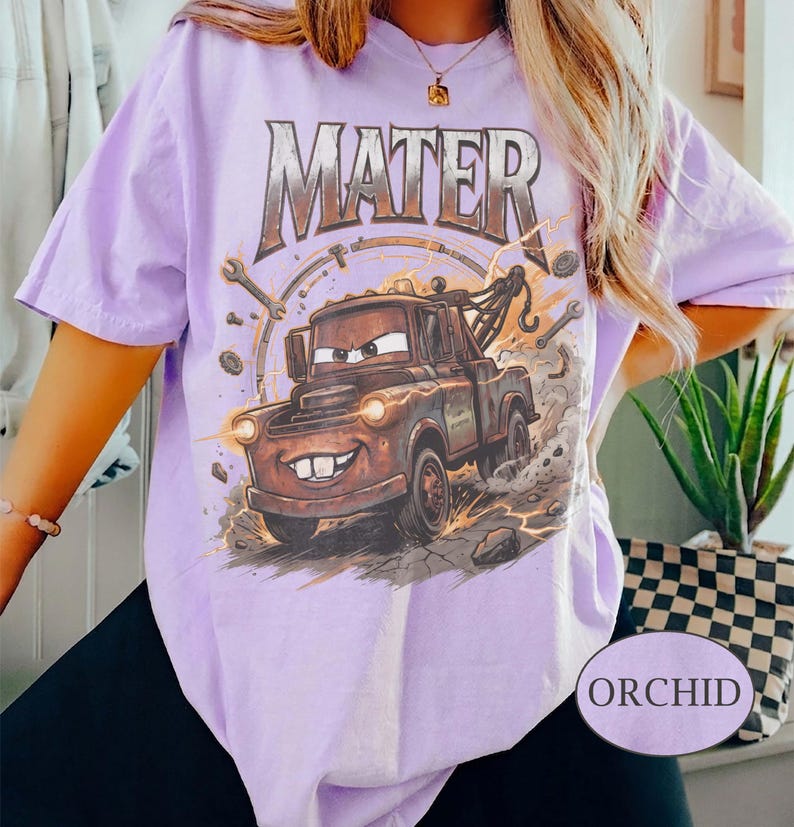 May include: A light purple t-shirt featuring a graphic print of a brown tow truck character with the text MATER in a metallic font above it. The design includes illustrations of wrenches, gears, and dust clouds surrounding the truck.