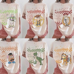 May include: A collection of six off-white t-shirts featuring Toy Story characters including Buzz Lightyear, Woody, Jessie, Mr. Potato Head, Rex, and Slinky. Each shirt displays a retro-style graphic with a checkered pattern and the character name.