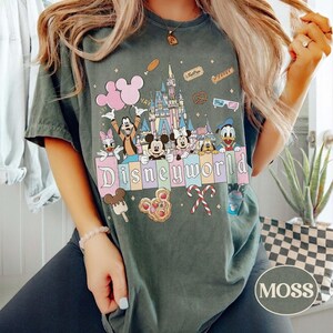 May include: Dark green t-shirt with a Disney World graphic. The design features cartoon characters, a castle, and the word "Disneyworld" in pastel colours. Additional graphics include balloons, ice cream, and candy canes.