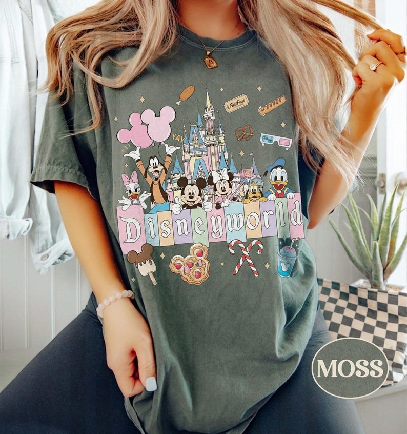 May include: Dark green t-shirt with a Disney World graphic. The design features cartoon characters, a castle, and the word "Disneyworld" in pastel colours. Additional graphics include balloons, ice cream, and candy canes.