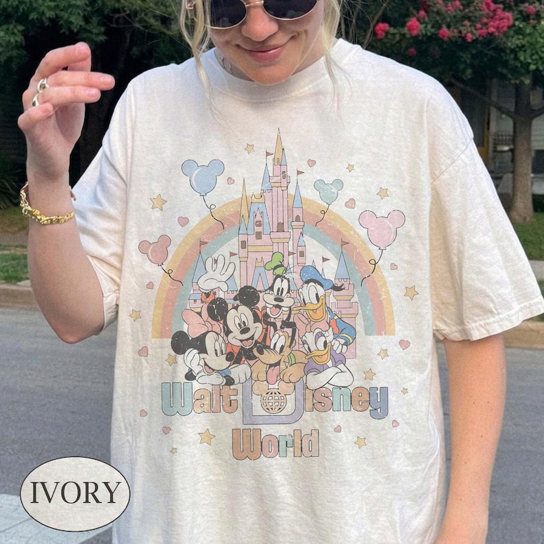 May include: Ivory-colored t-shirt featuring a colorful graphic of Disney characters, a castle, and a rainbow. The text "Walt Disney World" is printed below the graphic. The shirt has short sleeves and a relaxed fit.