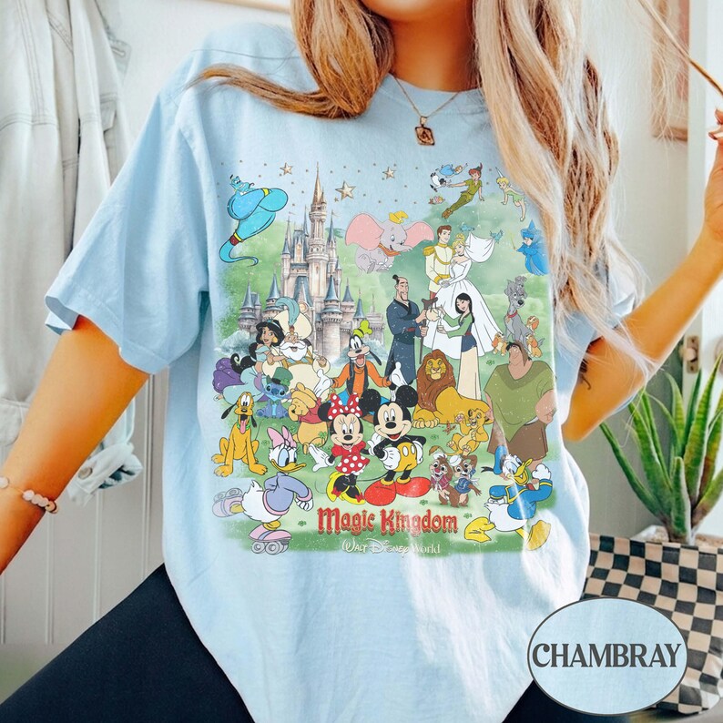 May include: Light blue t-shirt featuring a colorful graphic of Disney characters, including Mickey and Minnie Mouse, Aladdin, and Cinderella. The graphic includes the text "Magic Kingdom" and "Walt Disney World". The shirt has short sleeves and a relaxed fit.