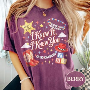 May include: A berry-colored t-shirt featuring a graphic print with the text I KNEW IT, I KNEW YOU, TOY STORY, INFINITY AND BEYOND, and YOU'VE GOT A FRIEND IN ME. The design includes a yellow sheriff star, red cowboy boots, a red hat, and alphabet blocks.