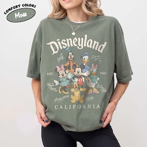 May include: A moss green t-shirt with the Disneyland logo and cartoon characters such as Mickey Mouse, Minnie Mouse, Donald Duck, and Goofy. The shirt also has the text "CALIFORNIA" and "EST. 1955".