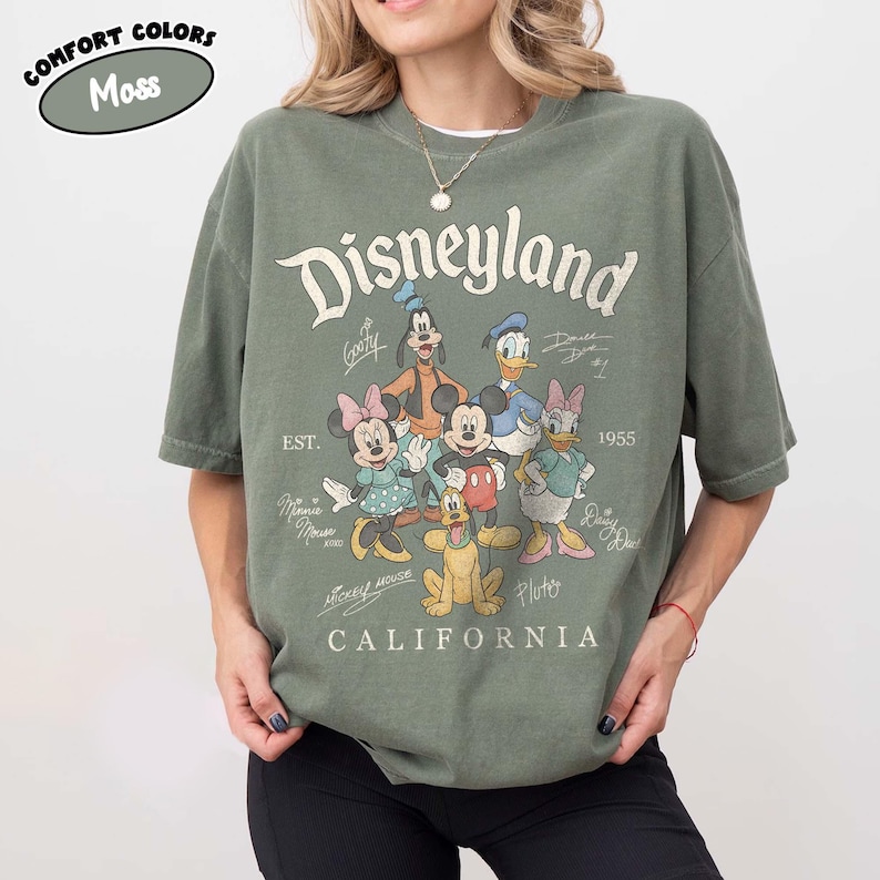 May include: A moss green t-shirt with the Disneyland logo and cartoon characters such as Mickey Mouse, Minnie Mouse, Donald Duck, and Goofy. The shirt also has the text "CALIFORNIA" and "EST. 1955".