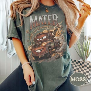 May include: A dark green short-sleeve t-shirt featuring a graphic print of the character Mater from the movie Cars. The design includes the text MATER in large letters above the rusty tow truck, surrounded by lightning bolts and mechanical tools.