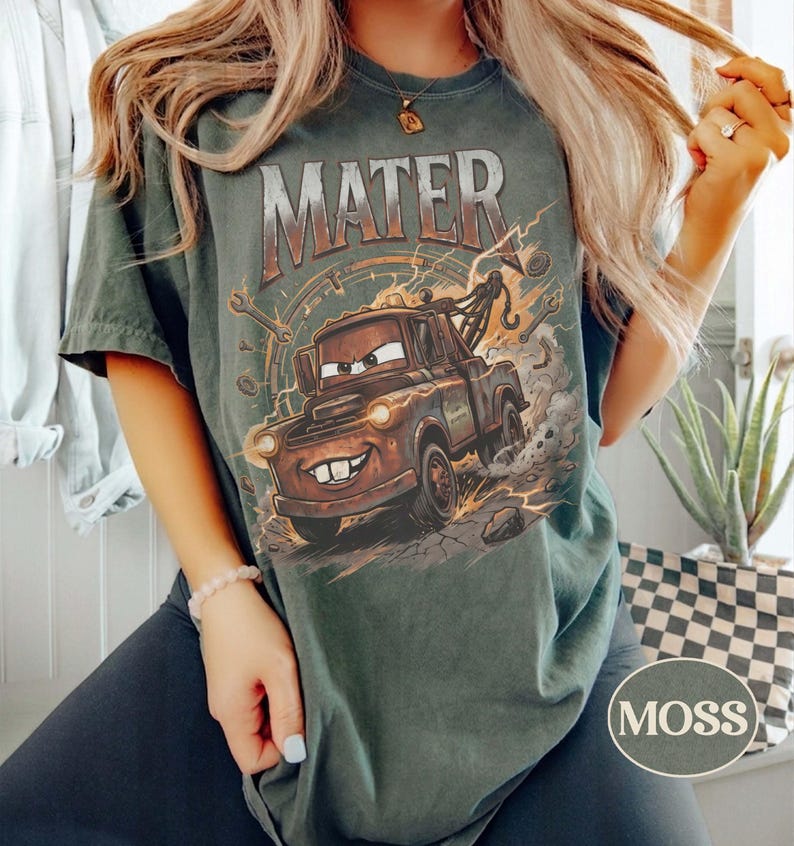 May include: A dark green short-sleeve t-shirt featuring a graphic print of the character Mater from the movie Cars. The design includes the text MATER in large letters above the rusty tow truck, surrounded by lightning bolts and mechanical tools.