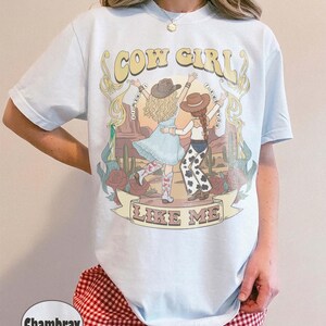 Puede incluir: Una camiseta azul claro con un estampado gr&aacute;fico de dos figuras con sombreros de vaquero en un paisaje des&eacute;rtico con cactus y rosas. El texto dice COW GIRL en la parte superior y LIKE ME en una pancarta en la parte inferior.