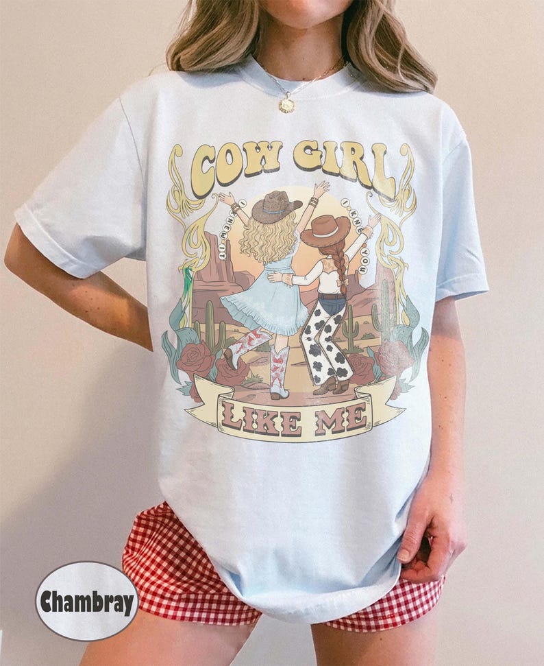 Puede incluir: Una camiseta azul claro con un estampado gr&aacute;fico de dos figuras con sombreros de vaquero en un paisaje des&eacute;rtico con cactus y rosas. El texto dice COW GIRL en la parte superior y LIKE ME en una pancarta en la parte inferior.