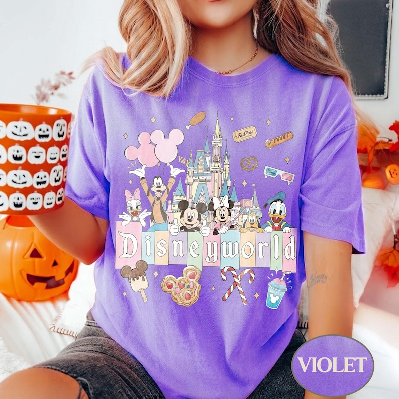May include: A violet t-shirt with a Disney World design. The graphic includes Mickey and Minnie Mouse, Donald Duck, and other characters, a castle, and food icons. The word "Disneyworld" is displayed in a colourful font.