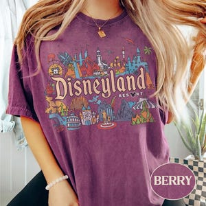 Op de afbeelding: Bessenkleurig T-shirt met een kleurrijke Disneyland Resort-afbeelding. De afbeelding bevat iconische bezienswaardigheden en het Disneyland Resort-logo. Het shirt heeft korte mouwen en een ronde hals.