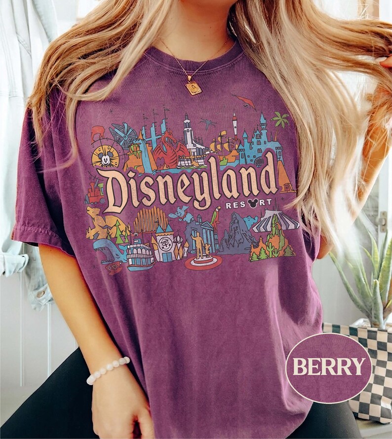 Op de afbeelding: Bessenkleurig T-shirt met een kleurrijke Disneyland Resort-afbeelding. De afbeelding bevat iconische bezienswaardigheden en het Disneyland Resort-logo. Het shirt heeft korte mouwen en een ronde hals.