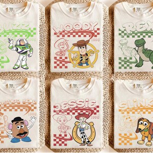 May include: Six off-white t-shirts featuring Toy Story characters: Buzz Lightyear, Woody, Rex, Mr. Potato Head, Jessie, and Slinky Dog. Each shirt displays the character name, a checkered pattern, the year 1995, and a line-art sketch of the character.