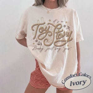 May include: An ivory short-sleeve t-shirt featuring a brown rope-style graphic that reads "it's a Toy Story" with the text "Baby, just say 'yes'" below it. The design includes illustrations of cacti, small flowers, and stars in a muted brown tone.