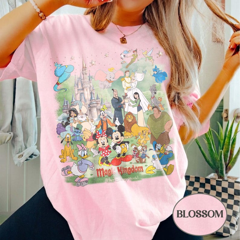 May include: Light pink t-shirt featuring a colorful graphic of Disney characters and a castle. The graphic includes Mickey and Minnie Mouse, Donald Duck, and other beloved characters. The text "Magic Kingdom" is also visible.