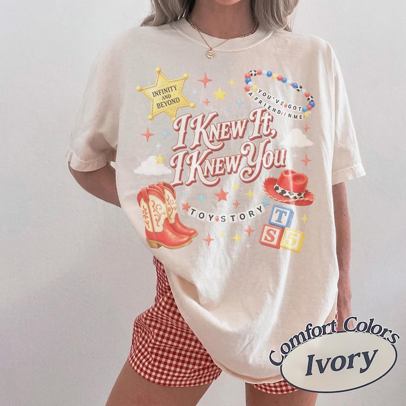 May include: An ivory short-sleeve t-shirt featuring a graphic print with the text "I KNEW IT, I KNEW YOU", "INFINITY AND BEYOND", "YOU'VE GOT A FRIEND IN ME", "TOY STORY", and "T S". The design includes a yellow star, red cowboy boots, a red hat, and blocks.