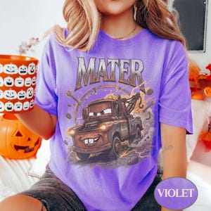 May include: A violet short-sleeve t-shirt featuring a graphic print of the character Mater from the movie Cars. The design includes the text MATER in a metallic font above the brown tow truck, surrounded by mechanical tools and lightning bolts.
