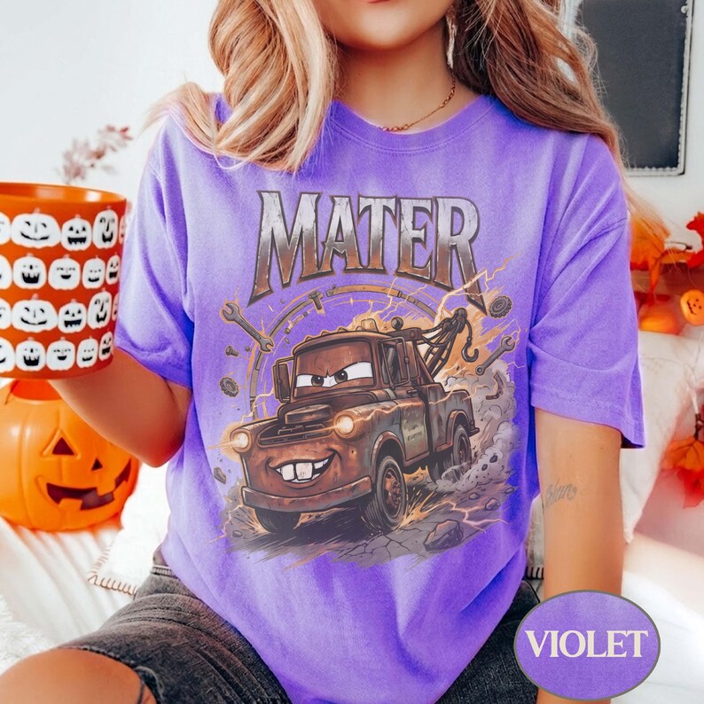 May include: A violet short-sleeve t-shirt featuring a graphic print of the character Mater from the movie Cars. The design includes the text MATER in a metallic font above the brown tow truck, surrounded by mechanical tools and lightning bolts.