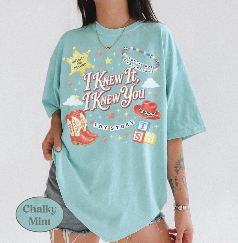 May include: A light mint green t-shirt featuring a graphic print with the text I KNEW IT, I KNEW YOU, TOY STORY, INFINITY AND BEYOND, and YOU'VE GOT A FRIEND IN ME. The design includes a yellow star, a beaded necklace, a red cowboy hat, boots, and blocks.
