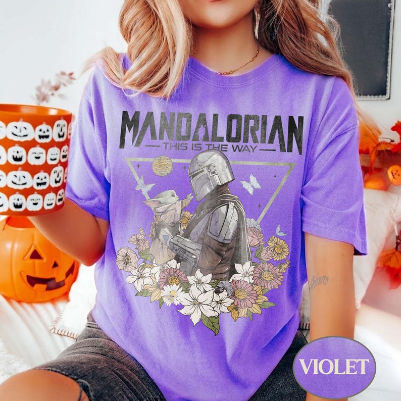 Op de afbeelding: Violet T-shirt met een afbeelding van The Mandalorian en Grogu, omringd door bloemen. De tekst "MANDALORIAN - THIS IS THE WAY" staat boven de afbeelding. Een Halloween-mok met pompoengezichten wordt in de linkerhand gehouden.