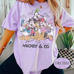 K&ouml;nnte beinhalten: Ein helllila T-Shirt mit einer Grafik von Mickey Mouse und seinen Freunden, darunter Donald Duck, Goofy, Minnie Mouse und Pluto. Das Shirt hat den Text "Mickey & Co" und "Est. 1928". Das Shirt ist mit "Orchid" beschriftet.