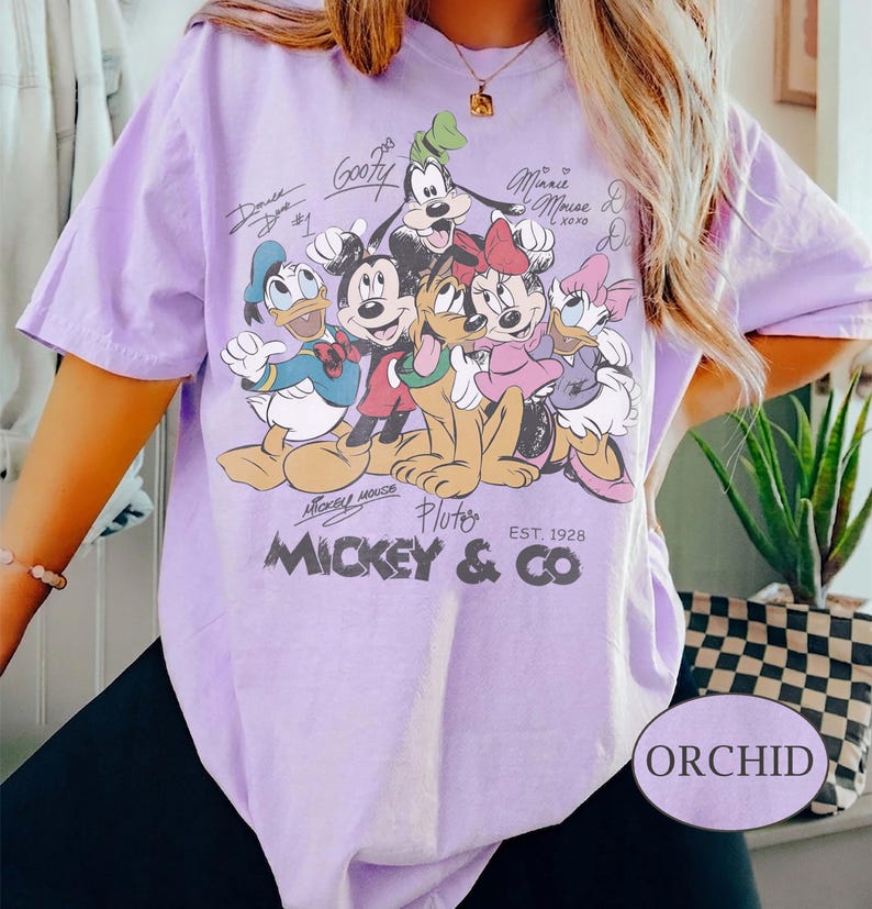 K&ouml;nnte beinhalten: Ein helllila T-Shirt mit einer Grafik von Mickey Mouse und seinen Freunden, darunter Donald Duck, Goofy, Minnie Mouse und Pluto. Das Shirt hat den Text "Mickey & Co" und "Est. 1928". Das Shirt ist mit "Orchid" beschriftet.