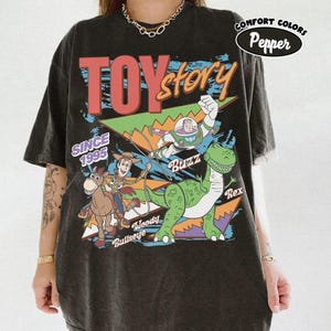 May include: Black t-shirt featuring a colorful graphic of Toy Story characters, including Woody, Buzz Lightyear, and Rex. The shirt has the text "TOY STORY" and "SINCE 1995". The shirt is from Comfort Colors.
