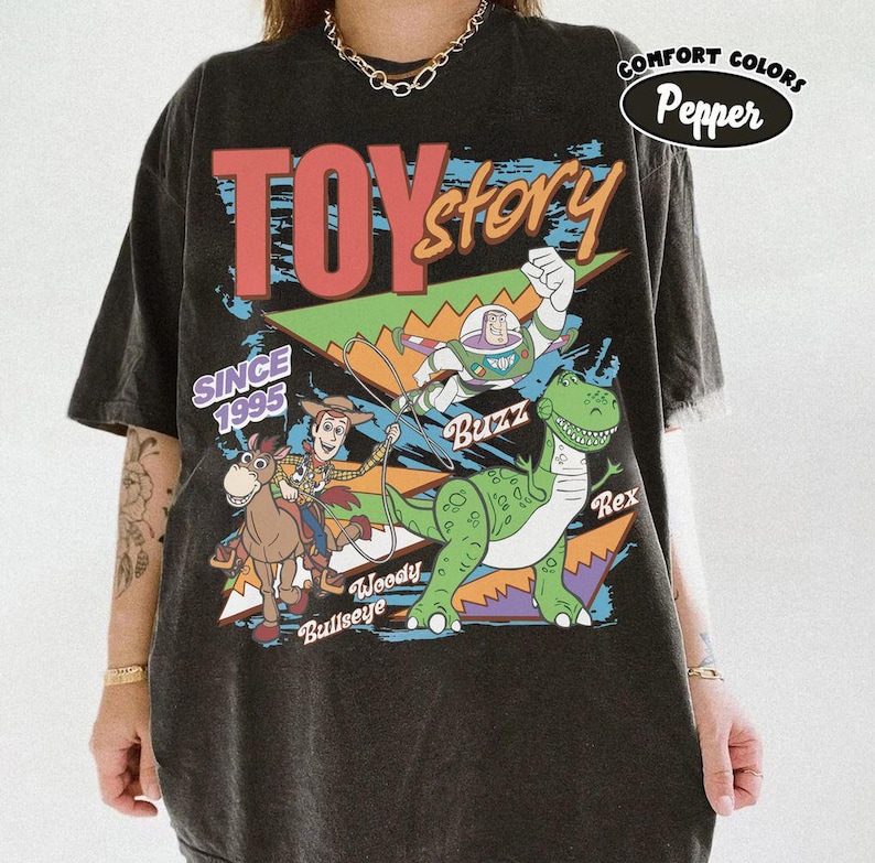 May include: Black t-shirt featuring a colorful graphic of Toy Story characters, including Woody, Buzz Lightyear, and Rex. The shirt has the text "TOY STORY" and "SINCE 1995". The shirt is from Comfort Colors.
