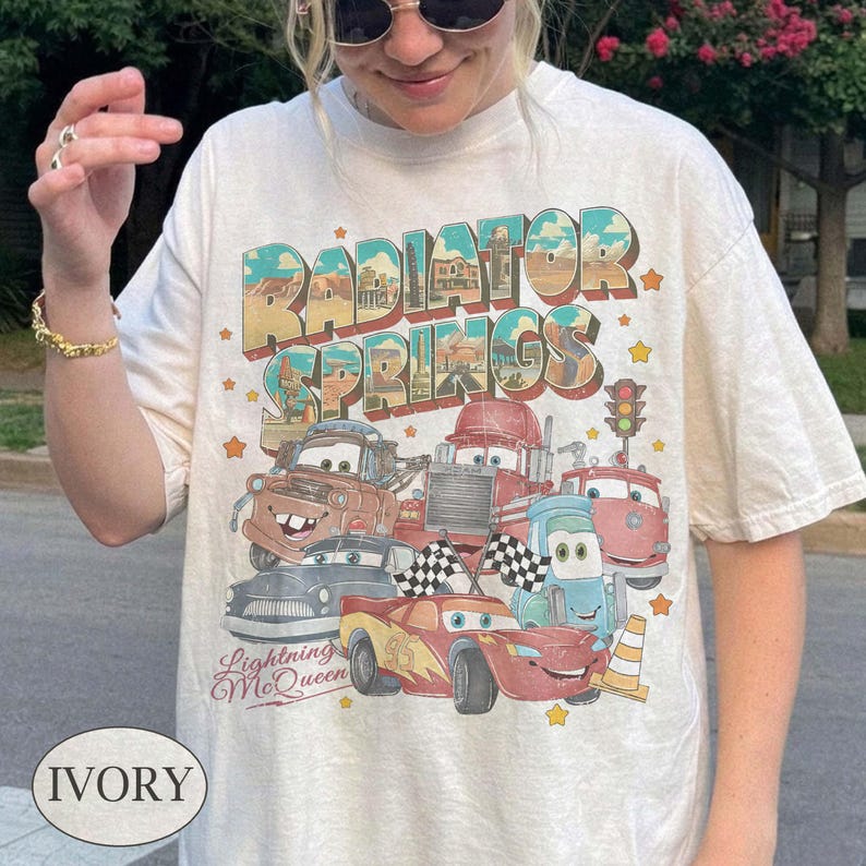 K&ouml;nnte beinhalten: Elfenbeinfarbenes T-Shirt mit einem farbenfrohen Aufdruck von Zeichentrickfiguren und dem Text "Radiator Springs" und "Lightning McQueen". Das Design umfasst verschiedene Zeichentrickautos, Sterne und eine Ampel.