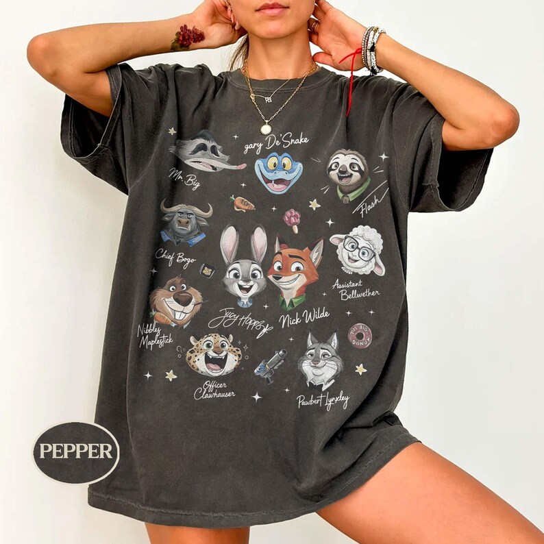 May include: A charcoal gray short-sleeve t-shirt featuring a graphic print of various animated animal characters from Zootopia, including Judy Hopps, Nick Wilde, and Chief Bogo, with their names printed in white script text alongside small star icons.
