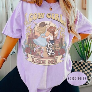 Puede incluir: Una camiseta de color morado claro con un gr&aacute;fico de dos figuras con sombreros de vaquero y botas en un paisaje des&eacute;rtico con cactus y rosas. El texto dice COW GIRL LIKE ME, con el texto I KNEW YOU alrededor.