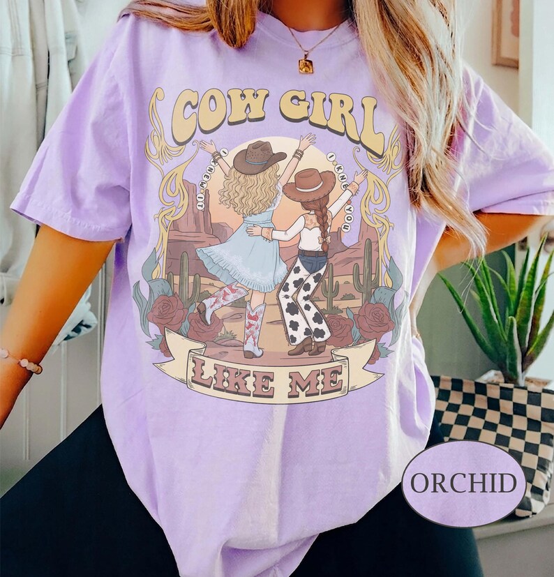 Puede incluir: Una camiseta de color morado claro con un gr&aacute;fico de dos figuras con sombreros de vaquero y botas en un paisaje des&eacute;rtico con cactus y rosas. El texto dice COW GIRL LIKE ME, con el texto I KNEW YOU alrededor.