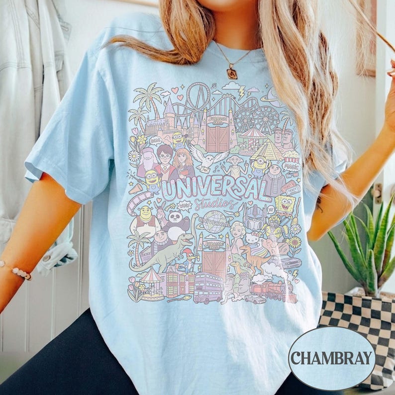 K&ouml;nnte beinhalten: Hellblaues T-Shirt mit einem farbenfrohen Grafikdesign von Universal Studios-Charakteren und -Attraktionen. Das Design enth&auml;lt den Text "UNIVERSAL Studios" in einer verspielten Schriftart, umgeben von Cartoon-Illustrationen.